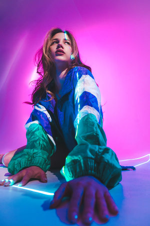 Young woman with long hair wears a colorful windbreaker, sitting against a vibrant pink background...