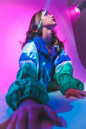 Young woman with long hair wears a colorful windbreaker, sitting against a vibrant pink background...
