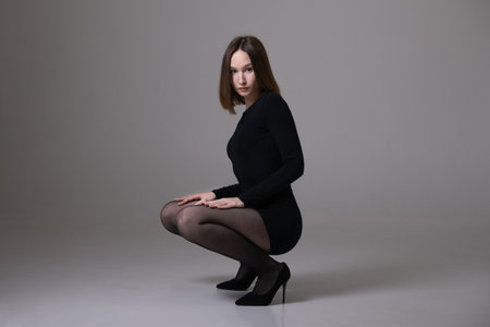 Young elegant woman in a black dress and tights crouches on heels on photo studioの写真素材