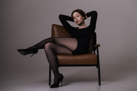 A young woman in a black outfit and heels poses confidently while sitting on a leather chair in a minimalist photo studio.の写真素材