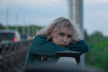 A woman green dress leans on a city bridge railing at dusk. Girl looking sad and lost in thought,...