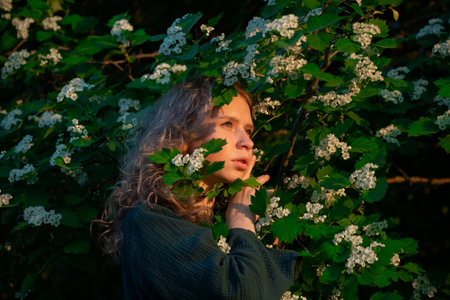 Young woman with curly blonde hair touches white flowers on a green bush in a forest, wearing a dark...