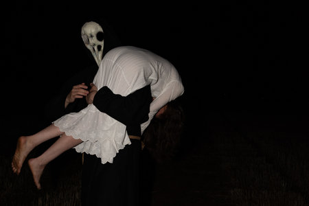 A man in a black robe and a raven skeleton mask holds a bound girl in a white lace dress on his shoulder at night in a rural field.の写真素材