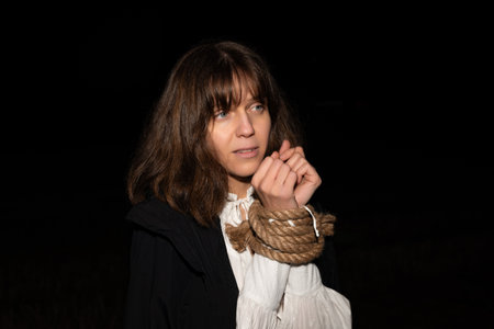 A young woman with brown hair and a white dress stands outside at night with hands tied by rope,...