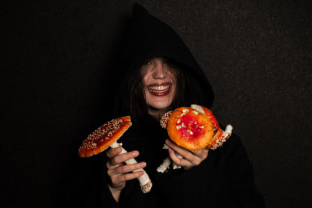 Witch woman dressed with hood in dark clothing holds large, amanita mushrooms. Sorceress, laughing and intoxicated with fly agarics against a dark background.の写真素材