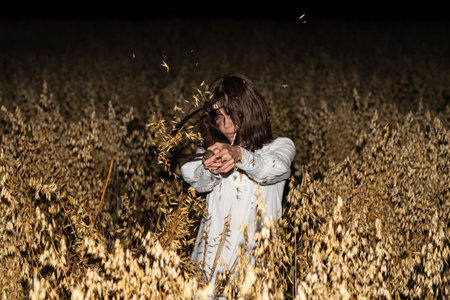 Woman in a white dress holding a sickle stands in a field of dry oats at night, with a menacing and eerie expression.の写真素材