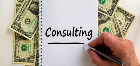 Male hand writing 'consulting' on white note, on white background. Dollar bills. Business concept, copy space.の写真素材