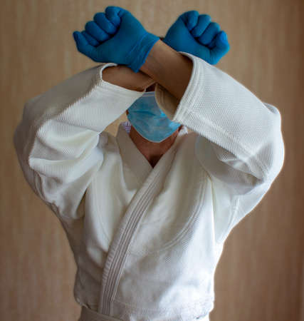 COVID-19 Pandemic Coronavirus concept. A young strong man in a white kimono for sambo, jiu jitsu and other martial arts with a blue medical gloves and medical mask.の写真素材