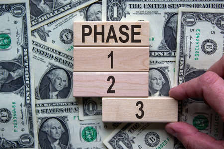 Wooden blocks form the words 'phase, 1, 2, 3,' on background from dollar bill. Male hand. Beautiful background. Business concept.の写真素材
