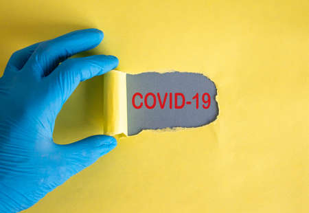The text covid-19 appearing behind torn yellow paper. Hand in blue glove.の写真素材