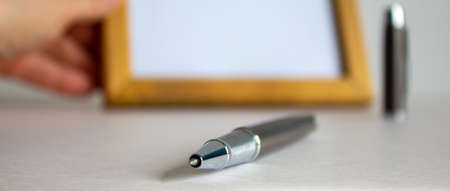 Pen on beautiful white fon. Wooden picture frame, male hand and cap in the background.の写真素材