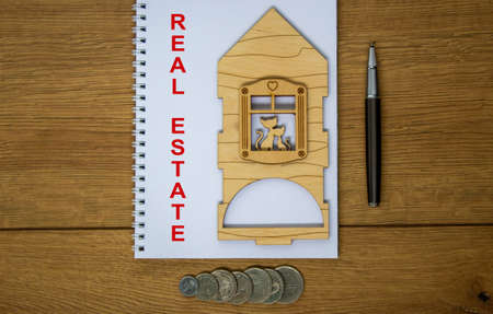 White note with inscription 'real estate' on beautiful wooden table, metalic pen, coins and miniature wooden house. Business concept.の写真素材