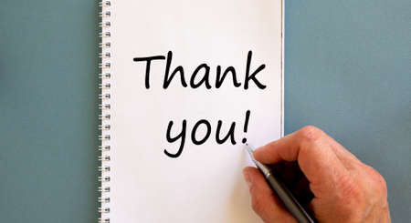 Male hand writes the words 'Thank you' on a white notebook. Beautiful blue background. Business concept.の写真素材