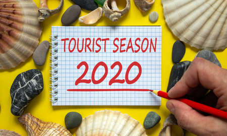 Male hand with red pencil writing 'tourist season 2020' on white note. Beautiful yellow background....