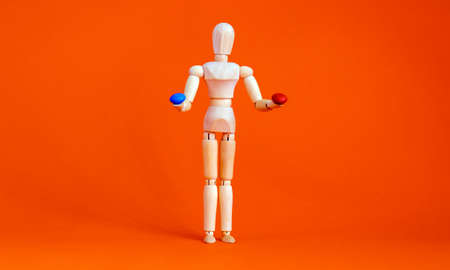 Wooden figure of a mannequin holding a red pill and blue pill on a orange background. Coronavirus,...