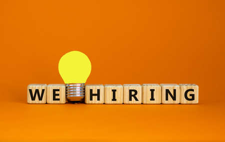 Wooden cubes with words 'we hiring'. Yellow light bulb. Beautiful orange background. Business concept. Copy space.の写真素材