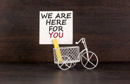 Wooden clothespin with white sheet of paper. Text 'we are here for you'. Miniature bicycle model. Beautiful dark wooden background. Business concept, copy space.の写真素材