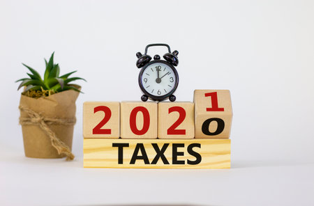Business and new year concept of taxes planning 2021. Fliped wooden cube and changed words 'TAXES 2020' to 'TAXES 2021'. Black alarm clock and house plant. Beautiful white background, copy space.の写真素材