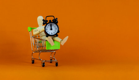 Sale time symbol. Wooden model of a human in a shopping cart with black alarm clock. Beautiful orange background. Business and sale concept, copy space.の写真素材