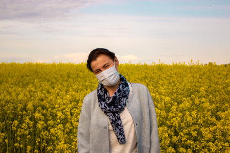 Woman in a medical mask on a background of a yellow flower field. Medical and COVID-19 Pandemic Coronavirus concept. Sky with clouds and sun.の写真素材