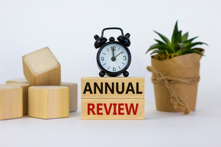 Annual review symbol. Concept words 'Annual review' on wooden blocks on a beautiful white background. Black alarm clock and house plant. Business and annual review concept.の写真素材