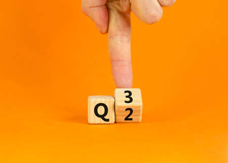 From 2nd to 3rd quarter symbol. Businessman turns a wooden cube and changes words 'Q2' to 'Q3'. Beautiful orange table, orange background. Business, happy 3rd quarter Q3 concept, copy space.の写真素材