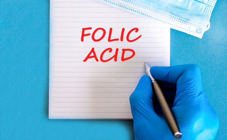 Folic acid symbol. Concept words Folic acid on beautiful white note. Doctor's hand in blue medical glove. Medical mask. Beautiful blue background. Medical healthy lifestyle folic acid concept.の写真素材