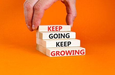 Keep going and growing symbol. Concept words phrase Keep going keep growing on beautiful wooden blocks. Beautiful orange background. Businessman's hand. Business Keep going growing concept. Copy spaceの写真素材