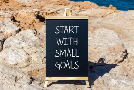 Start with small goals symbol. Concept words phrase Start with small goals on beautiful black blackboard. Beautiful stone blue sea sky background. Business Start with small goals concept. Copy space.の写真素材
