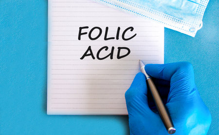 Folic acid symbol. Concept words Folic acid on beautiful white note. Doctor's hand in blue medical glove. Medical mask. Beautiful blue background. Medical healthy lifestyle folic acid concept.の写真素材