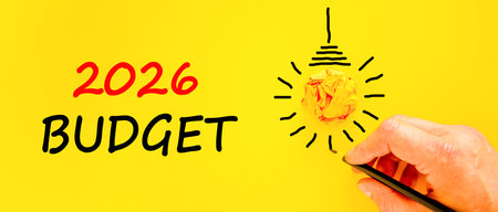 2026 Budget symbol. Concept words 2026 Budget on beautiful yellow paper. Beautiful orange paper background. Light bulb icon. Businessman's hand with pen. Business and 2026 budget concept. Copy space.の写真素材