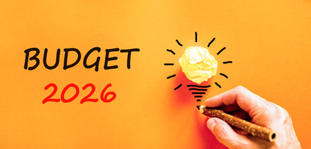 Budget 2026 symbol. Concept words Budget 2026 on beautiful orange paper. Beautiful orange paper background. Light bulb icon. Businessman's hand with pen. Business and budget 2026 concept. Copy space.の写真素材