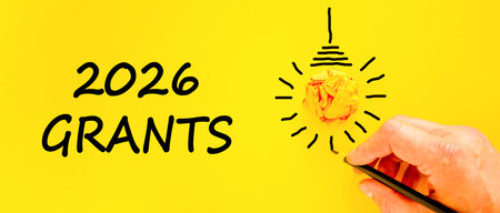2026 Grants symbol. Concept words 2026 Grants on beautiful yellow paper. Beautiful orange paper background. Light bulb icon. Businessman's hand with pen. Business and 2026 grants concept. Copy space.の写真素材