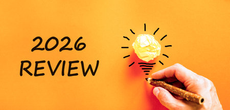 2026 Review symbol. Concept words 2026 Review on beautiful orange paper. Beautiful orange paper background. Light bulb icon. Businessman's hand with pen. Business and 2026 review concept. Copy space.の写真素材