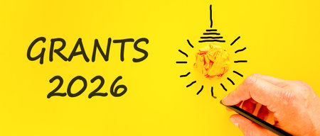Grants 2026 symbol. Concept words Grants 2026 on beautiful yellow paper. Beautiful orange paper background. Light bulb icon. Businessman's hand with pen. Business and grants 2026 concept. Copy space.の写真素材