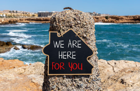 Support, help and we are here for you symbol. Concept words We are here for you on beautiful blackboard. Beautiful stone and blue sky, sea background. Business we are here for you concept. Copy space.の写真素材