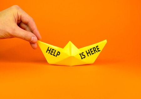 Help is here and support words symbol. Concept words Help is here on beautiful yellow paper boat. Beautiful orange background. Businessman hand. Business help is here and support concept. Copy space.