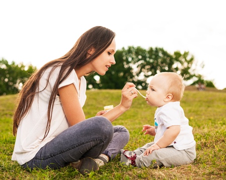 Yong  mother with long hair feeding her baby puree outdoors on grass  in sunlightの写真素材