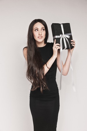 Young beautiful woman with long hair wearing black cocktail dress is holding elegantly wrapped gift.の写真素材