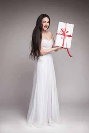 Young beautiful woman with long dark hair wearing maxi white dress, holding gift wrapped in white paper with red ribbon.の写真素材