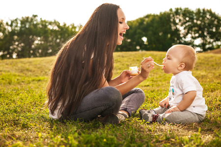 Young beautiful mother feeding her baby puree outdoors in sunlight