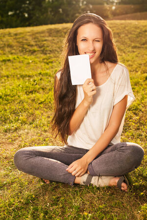 Closeup of young beautiful woman with long dark hair, holding blank white board on green background of summer natureの写真素材