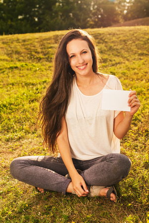 Young smiling beautiful woman with long dark hair is sitting on the grass, holding blank white boardの写真素材