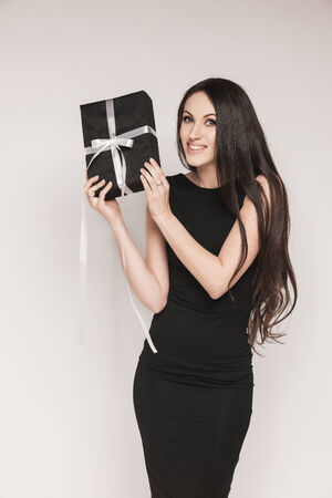 Young beautiful woman with long hair wearing black cocktail dress is holding elegantly wrapped gift.の写真素材