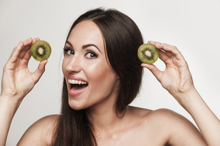 Funny portrait of young brunette woman holding sliced fresh kiwifruit. Healthy eating lifestyle and weight loss concept. Studio white background.の写真素材