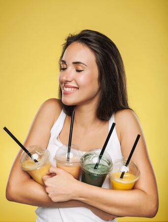 Young attractive brunette woman holding takeaway cups of smoothie. Happy girl posing on yellow background. Healthy eating concept.の写真素材