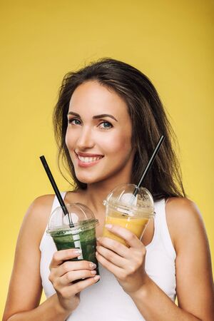 Young attractive brunette woman holding takeaway cups of smoothie. Happy girl posing on yellow background. Healthy eating concept.の写真素材