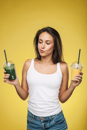 Young attractive brunette woman holding takeaway cups of smoothie. Happy girl posing on yellow background. Healthy eating concept.の写真素材