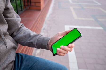 Young man in gray sweatshirt holds a phone with green screen outsideの写真素材