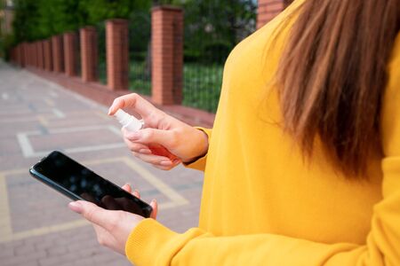 A young girl in a yellow sweater disinfects her phone with a spray-sanitizer on the streetの写真素材